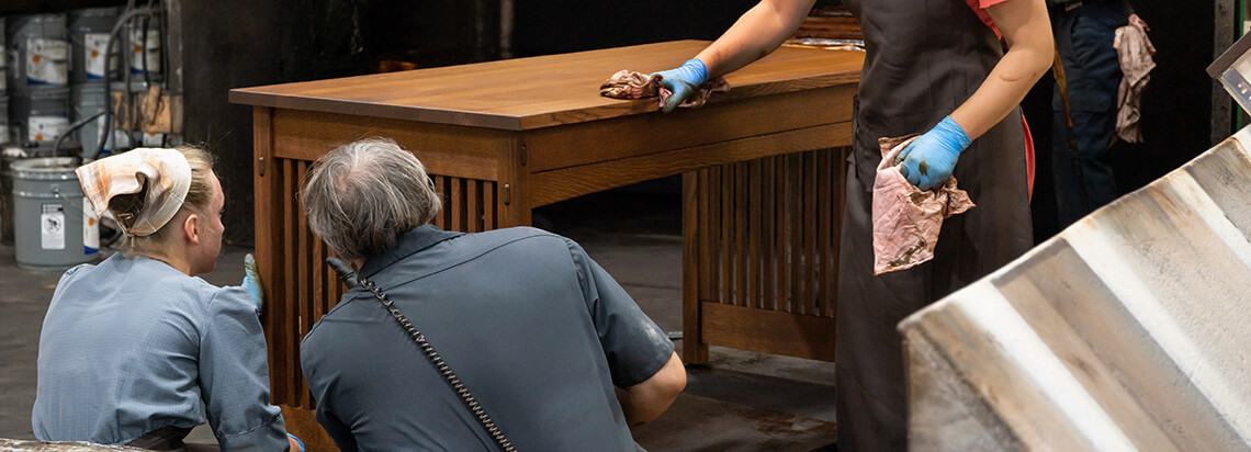 Amish furniture stain is applied to a quarter sawn white oak wood desk by Amish woodworkers in a finishing shop 